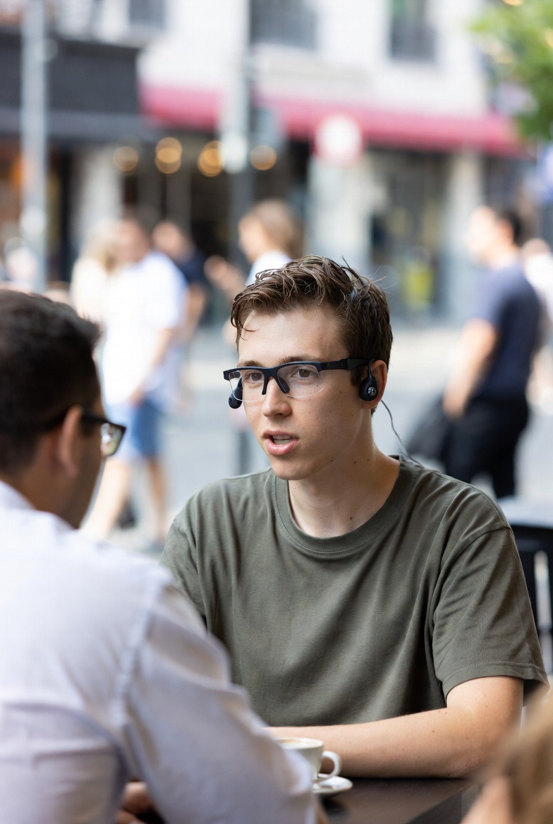 Person wearing smart glasses speaking with others at a cafe, small ear speakers visible